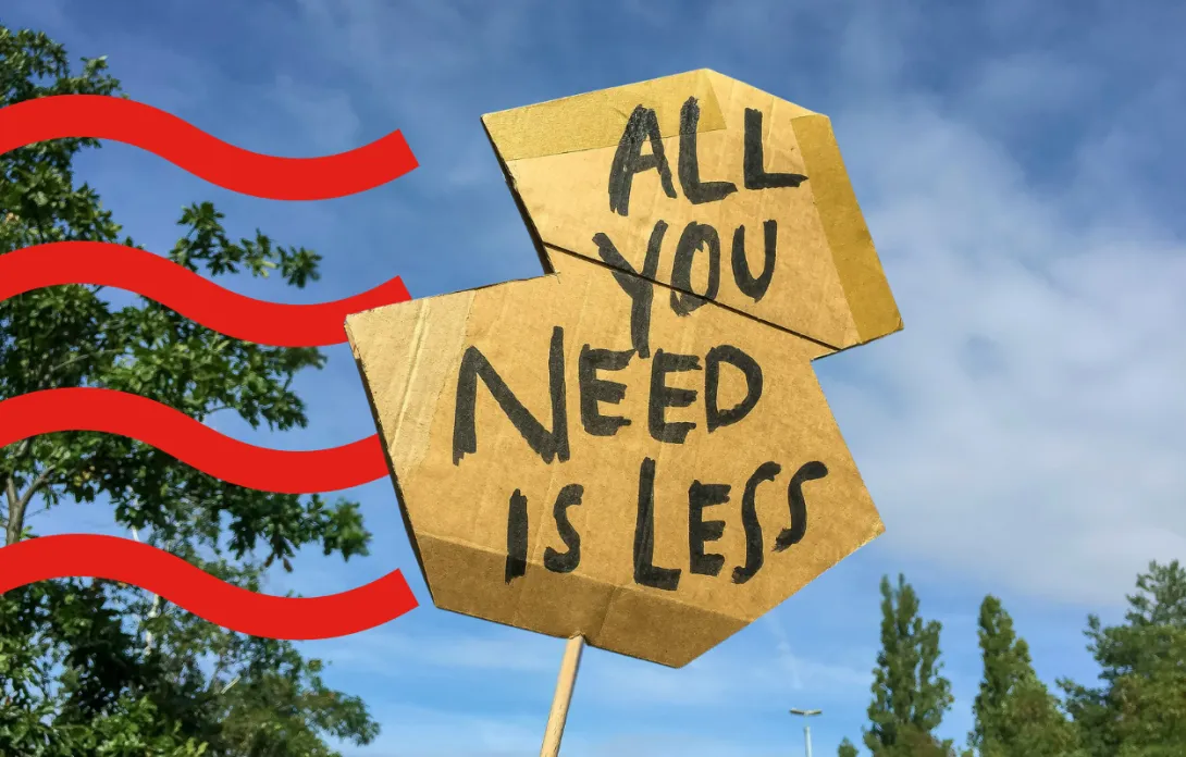 Cardboard protest sign reading ‘All you need is less’ held up outdoors.